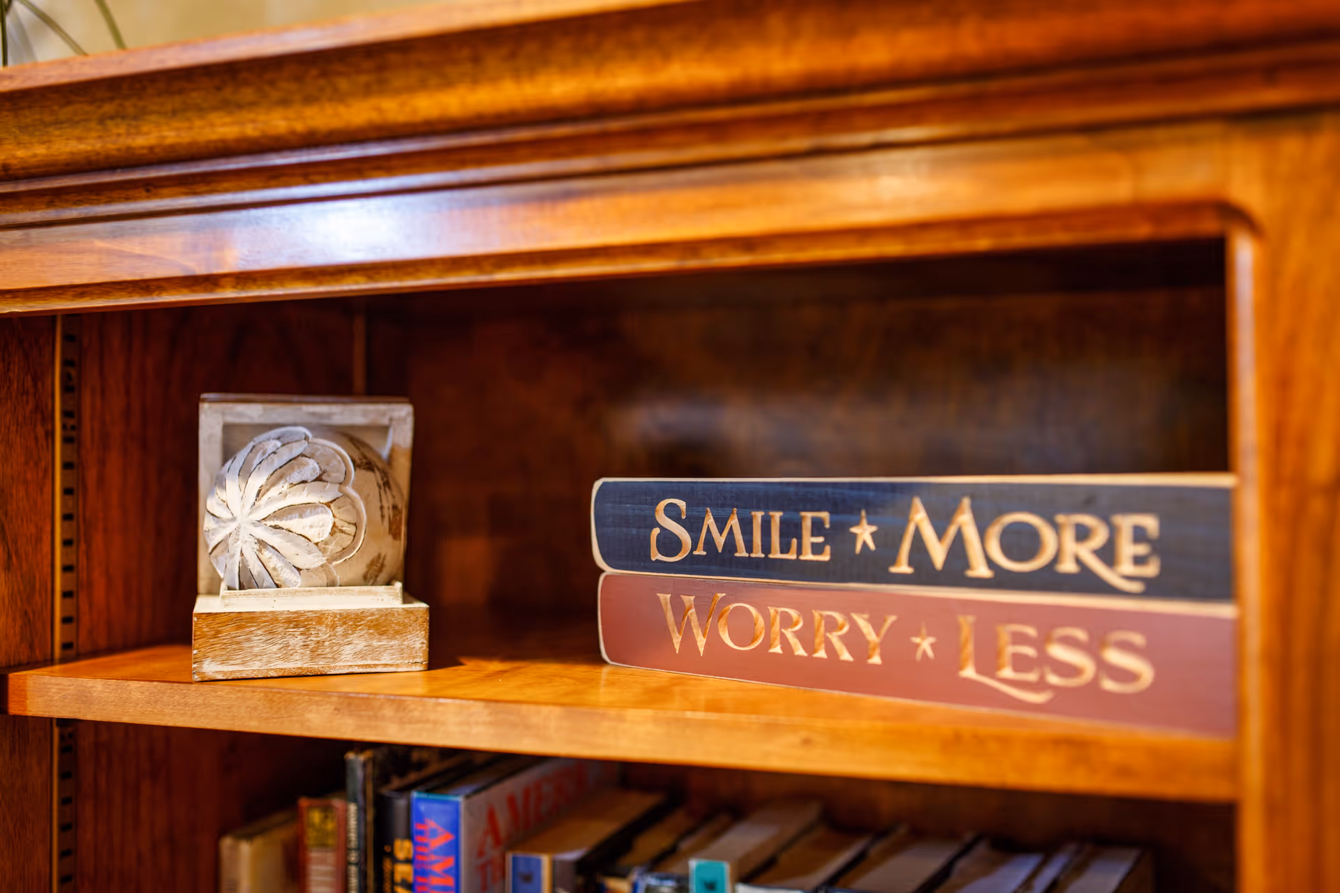 A wooden bookshelf with decorative items including a carved wooden block and two stacked wooden signs with the phrases 'Smile More' and 'Worry Less'. Below the shelf are several books arranged vertically.