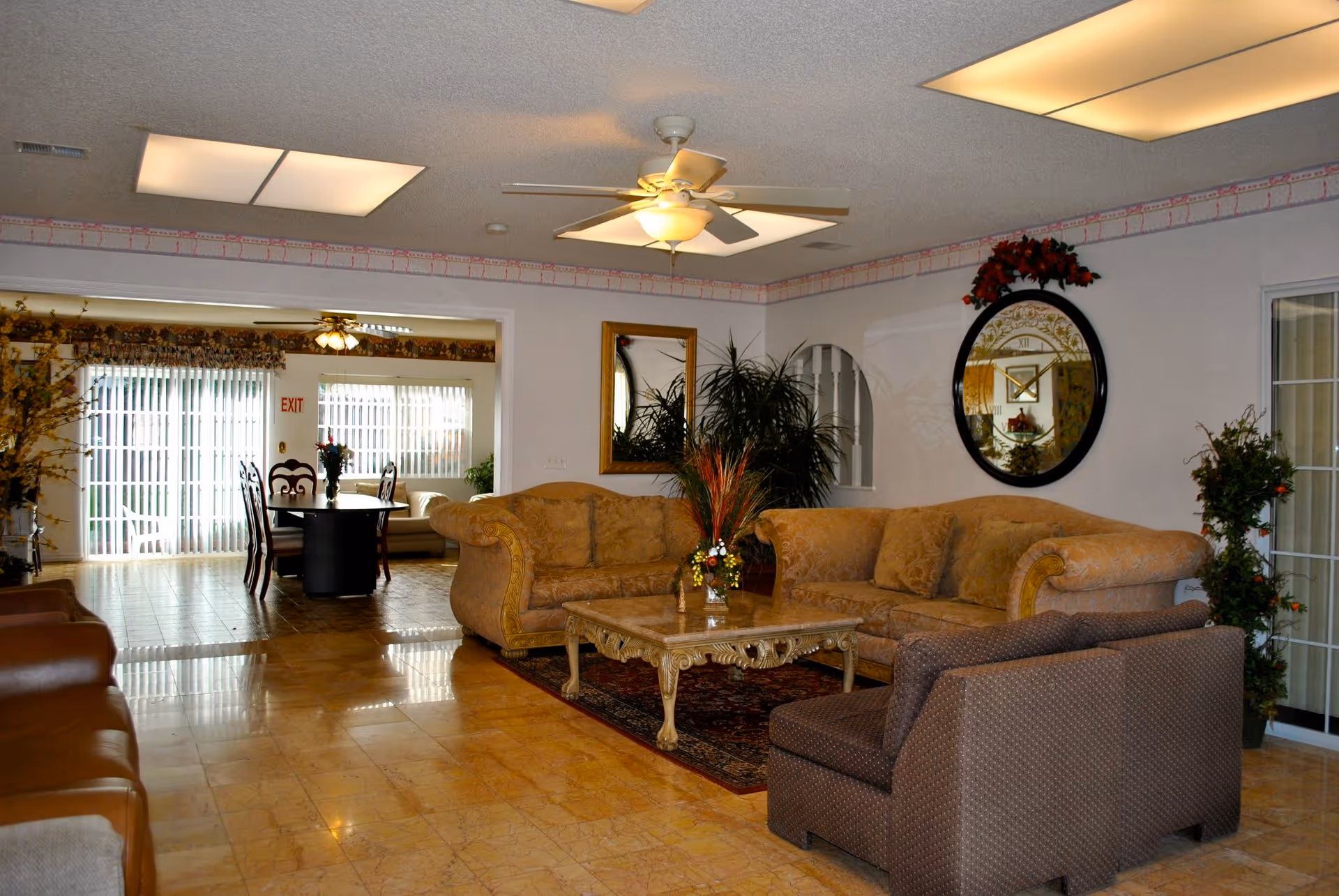 A cozy living room area with a beige patterned sectional sofa, a matching armchair, and a decorative coffee table on a rug. The room has a ceiling fan with lights, a large round wall clock, a rectangular mirror, and several potted plants. In the background, there is a dining area with a table and chairs near large windows with vertical blinds.