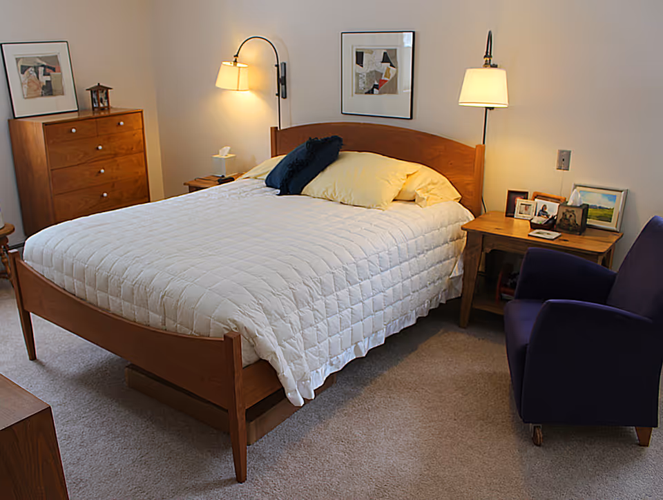 A cozy bedroom with a wooden bed frame and a white quilted bedspread. The bed has two pillows, one yellow and one dark blue. On either side of the bed are wooden nightstands with lamps mounted on the wall above them. The left nightstand holds a tissue box and a small item, while the right nightstand displays several framed photos and a tissue box. A wooden dresser with drawers and a framed picture on top is against the wall to the left. A dark blue armchair is positioned near the right nightstand. The room has beige carpeting and light-colored walls.