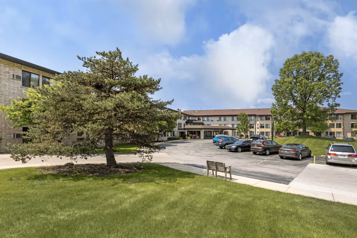 Exterior view of Oak Hill Terrace Senior Living facility showing a parking lot with several parked cars, a grassy area with a bench, and trees under a partly cloudy sky.