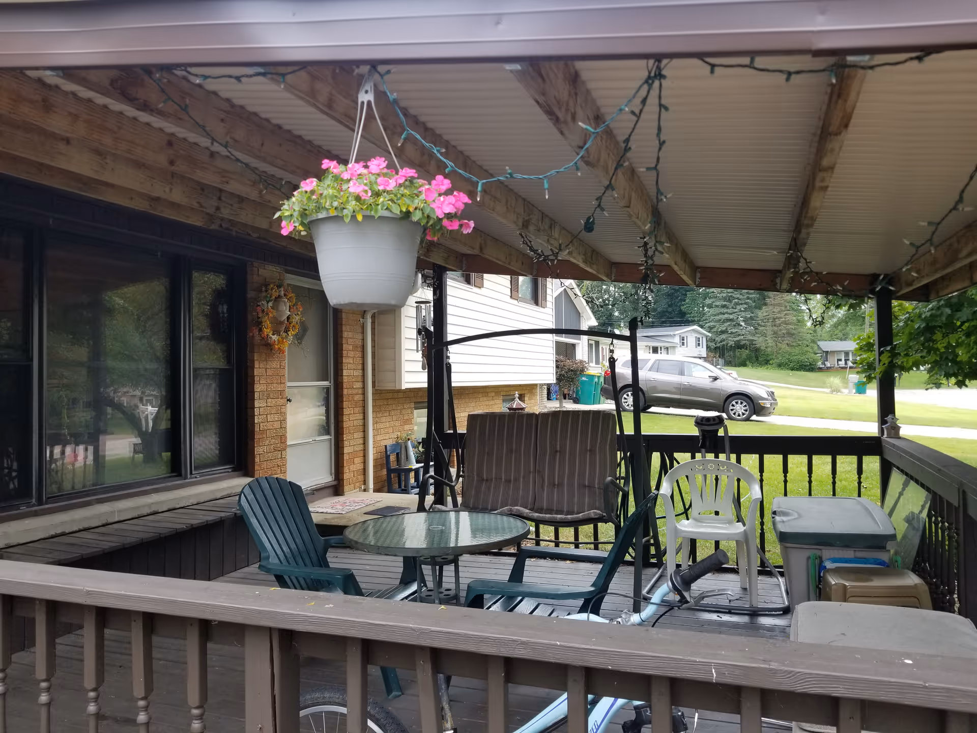 Covered outdoor patio area with wooden railing and ceiling beams. The patio has a hanging flower pot with pink flowers, a glass-top round table surrounded by plastic chairs, a cushioned swing seat, and various storage containers. In the background, a residential neighborhood with houses, a parked car, and green lawns is visible.