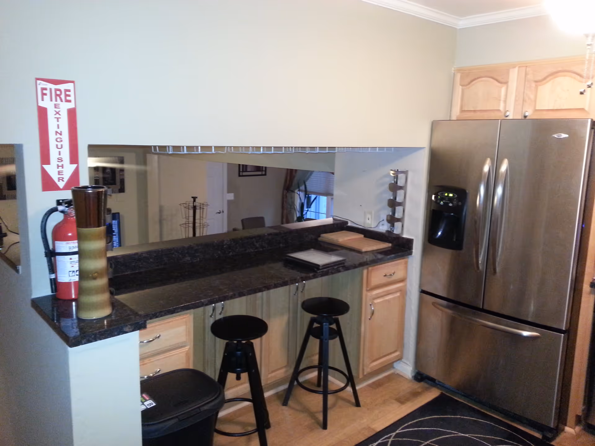 Interior view of a kitchen area featuring a stainless steel refrigerator with a water dispenser, light wooden cabinets, a black countertop with two black bar stools, a fire extinguisher mounted on the wall with a red and white sign above it, and a decorative vase on the counter.