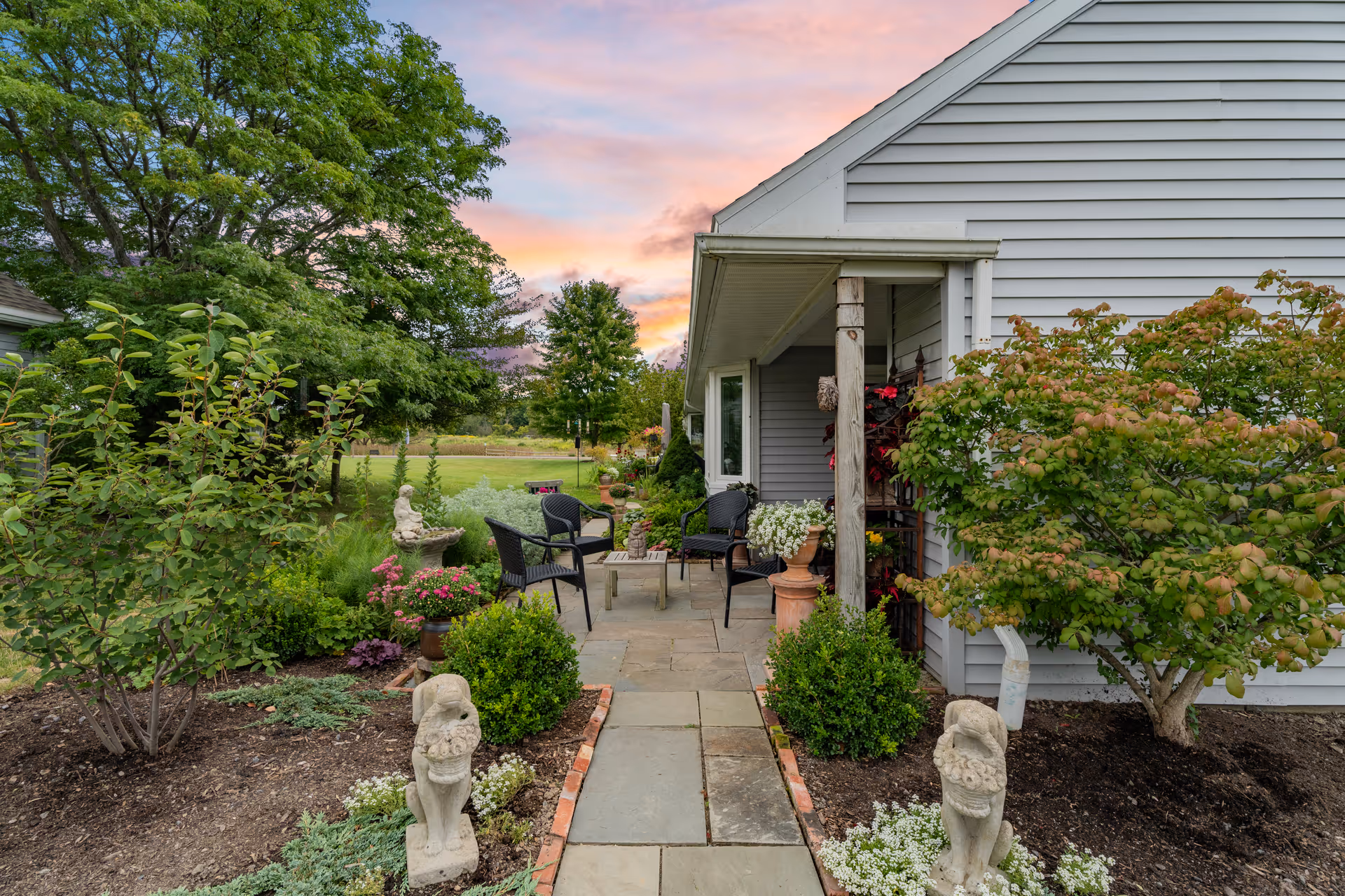 A peaceful outdoor patio area at Kendal at Ithaca with stone pathway leading to a seating area with black chairs and a small table. The patio is surrounded by lush greenery, flowering plants, and decorative stone statues, with a house exterior visible on the right and a colorful sunset sky in the background.
