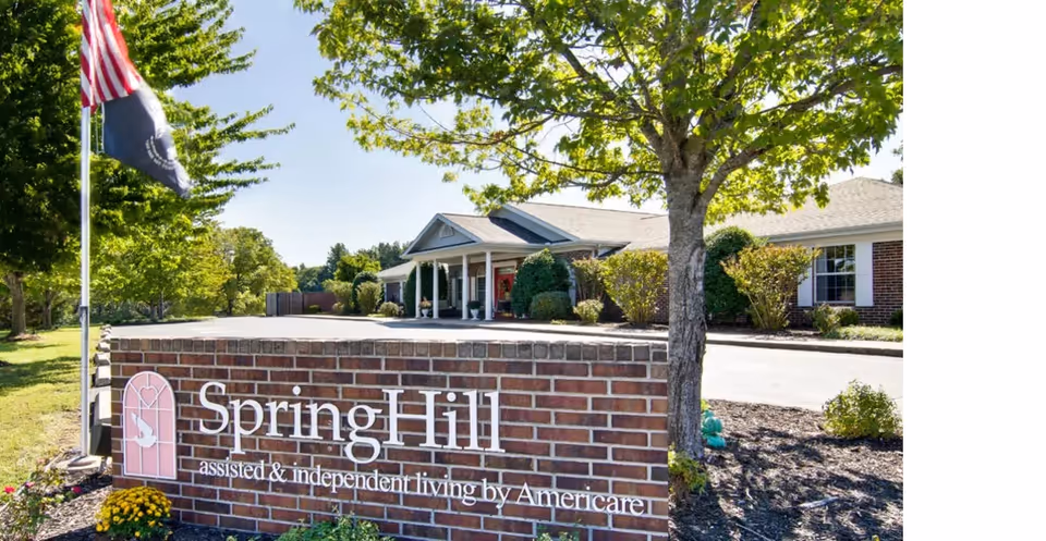 Exterior view of a senior living facility with a brick sign that reads 'SpringHill assisted & independent living by Americare'. The building is surrounded by trees and landscaping, with an American flag and another flag on a flagpole nearby.