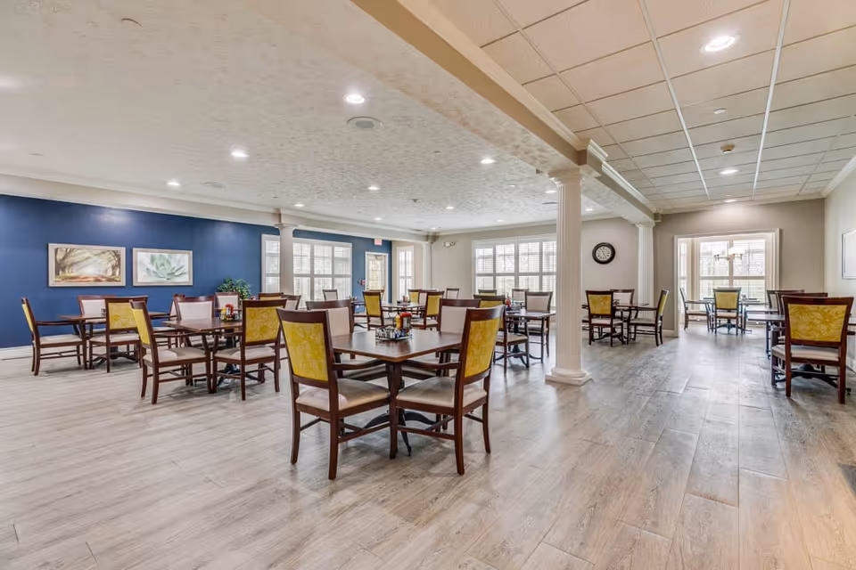 A spacious dining room in an assisted living facility with multiple wooden tables and chairs arranged neatly. The room features light wood flooring, a blue accent wall with framed artwork, large windows allowing natural light, and white columns supporting the ceiling.