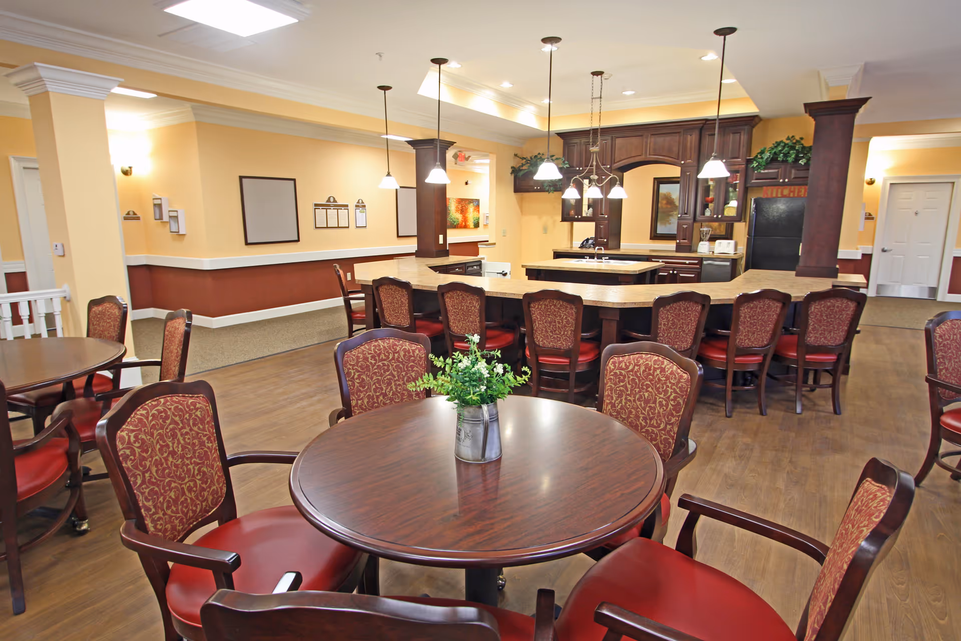 A spacious dining area with round wooden tables and upholstered chairs with red cushions and patterned backs. In the background, there is a kitchen area with a large island, dark wood cabinetry, pendant lighting, and a refrigerator. The walls are painted in warm tones with white trim, and the floor is wood.