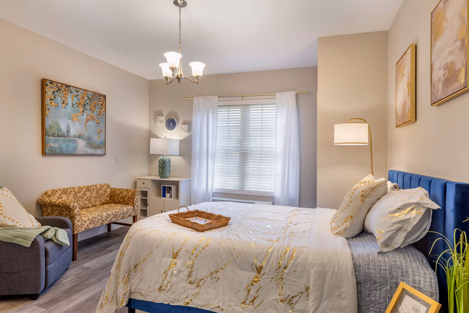A well-lit bedroom with a large bed featuring white and gold bedding, a blue upholstered headboard, and multiple pillows. There is a small wicker tray on the bed. To the left, there is a patterned loveseat and a gray armchair with a green blanket draped over it. A white cabinet with a green lamp and a wall clock is positioned against the wall near a window with white curtains. The walls are decorated with framed artwork, and a floor lamp stands beside the bed.