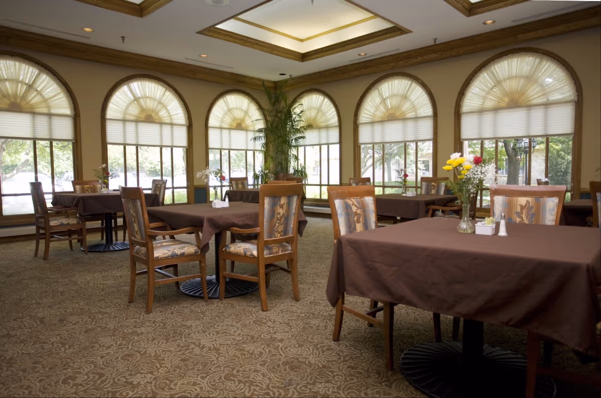 A dining room with several tables covered in brown tablecloths and wooden chairs with patterned cushions. Each table has a small vase with flowers and salt and pepper shakers. The room features large arched windows with pleated shades, allowing natural light to fill the space. There is a patterned carpet and a ceiling with recessed lighting.
