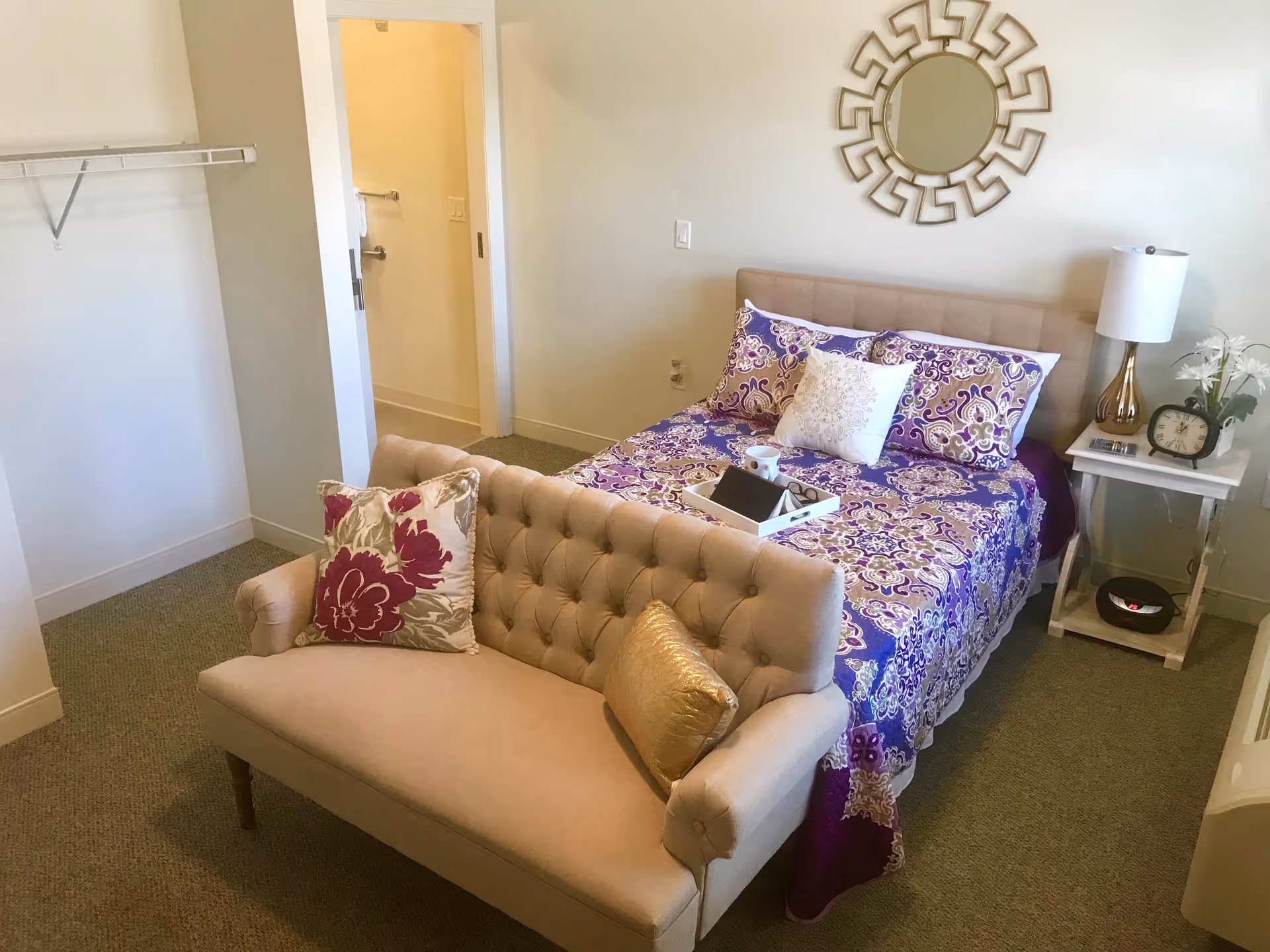 A cozy bedroom featuring a bed with purple and white patterned bedding, a beige tufted loveseat with decorative pillows at the foot of the bed, a white nightstand with a lamp, clock, and flowers, and a decorative round mirror on the wall. An open doorway leads to a bathroom area.
