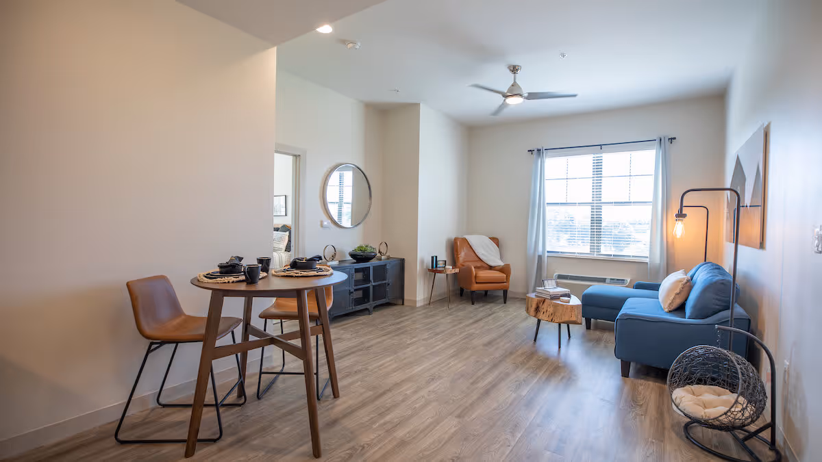 A bright and modern living room and dining area with light wood flooring. The dining area features a small round wooden table set for two with brown chairs. The living room has a blue sectional sofa, a wooden coffee table, a brown leather armchair, a floor lamp, and a hanging chair with a cushion. A large window with white curtains lets in natural light, and a ceiling fan is mounted above.