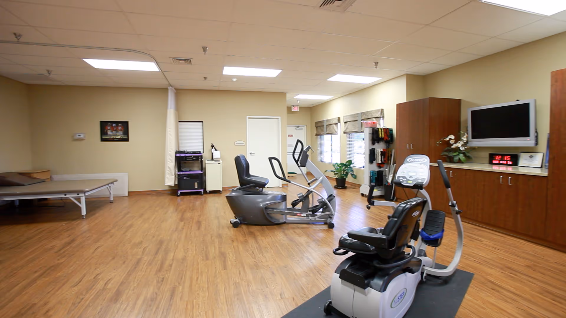 A spacious room with wooden flooring featuring exercise equipment including a recumbent bike and an elliptical machine. The room has beige walls, a treatment table on the left, a TV mounted on the right wall above wooden cabinets, and windows with blinds. There are also some plants and exercise accessories visible.