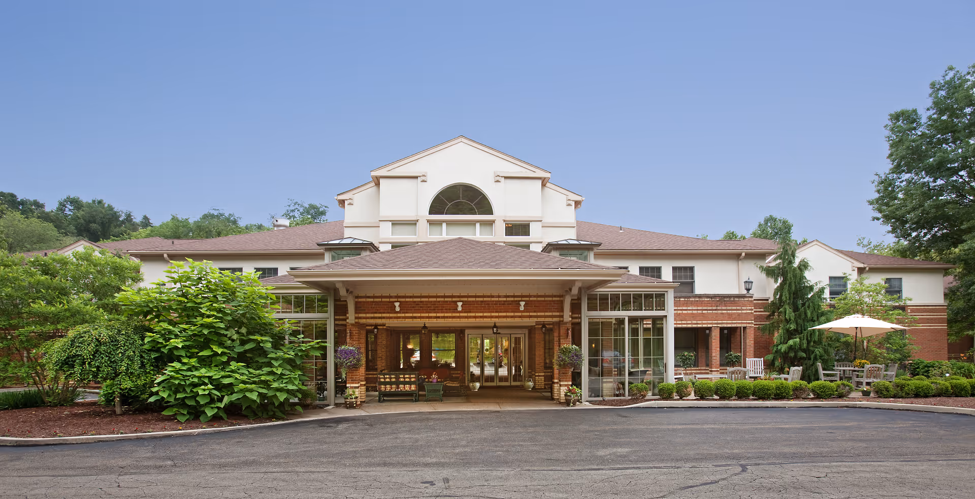 Front exterior of a senior living facility showing the main entrance with a covered porte-cochere, landscaped grounds, and outdoor seating.