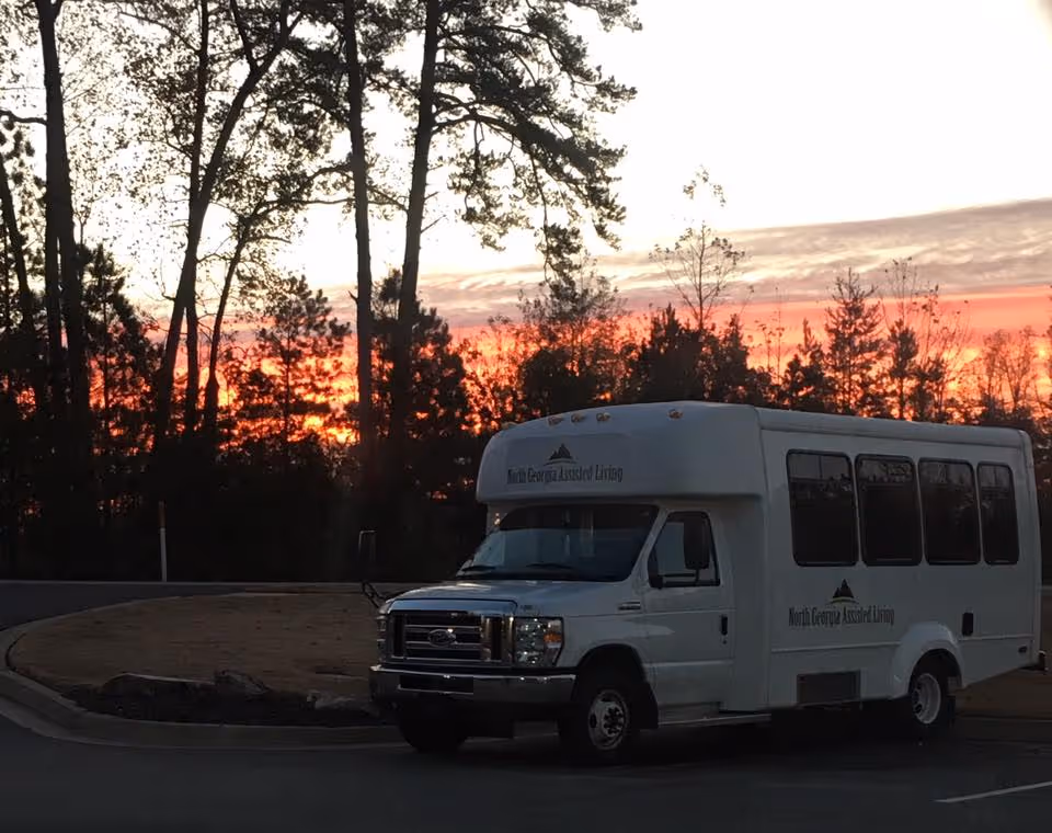 A white shuttle bus with the logo and text 'North Georgia Assisted Living' parked on a paved road near a wooded area during a vibrant sunset with orange and pink hues in the sky.