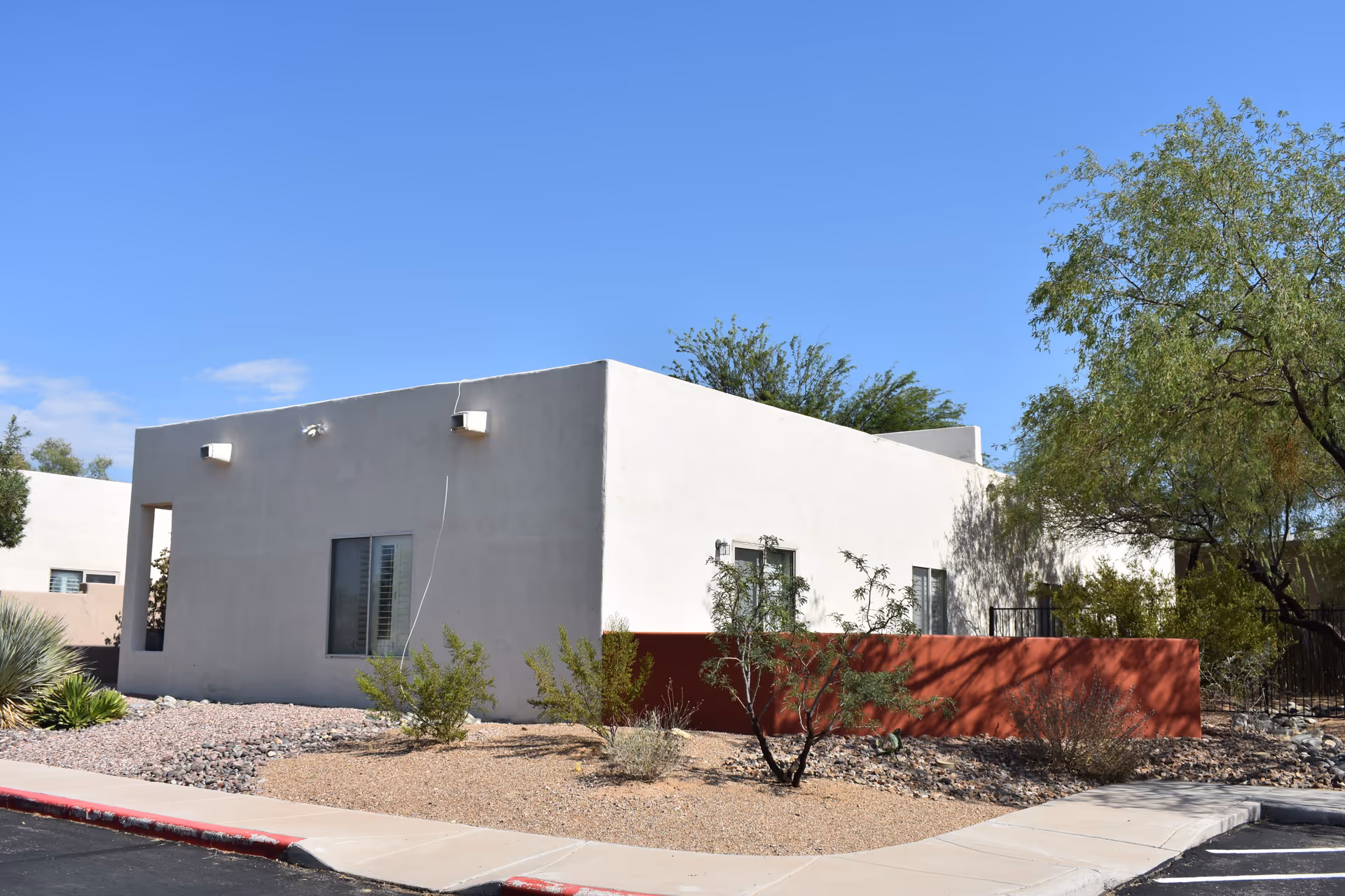 Exterior view of a single-story building with white walls and a red lower section, surrounded by desert landscaping with small bushes and trees under a clear blue sky.