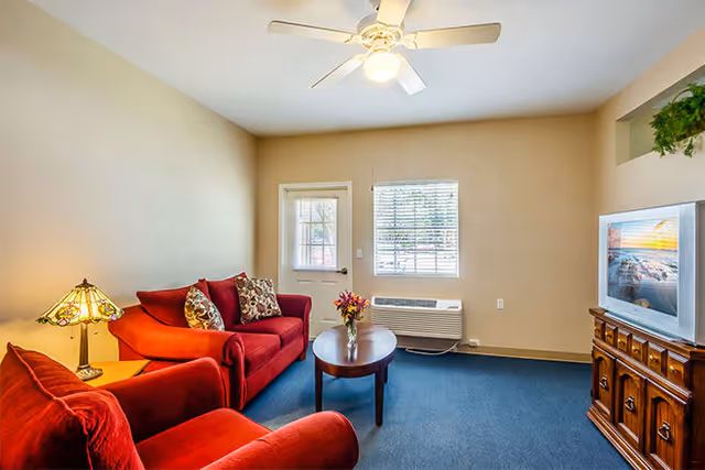 A cozy living room with a red sofa and armchair, a wooden coffee table with a flower vase, a TV on a wooden cabinet, a ceiling fan with light, a window with blinds, and a door leading outside.