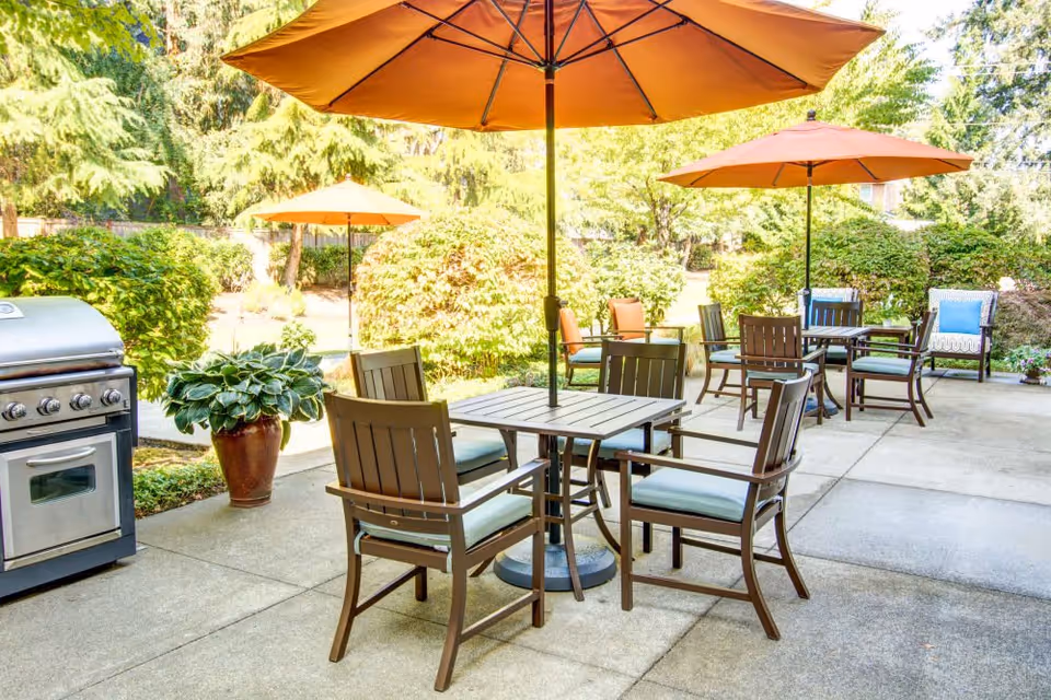 Outdoor patio area with several tables and chairs under orange umbrellas, surrounded by greenery and bushes. A stainless steel grill is visible on the left side, and the setting is bright and sunny.