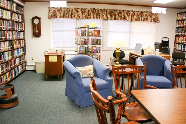 A cozy library room with bookshelves filled with books along the walls, two blue upholstered armchairs with a floral pillow on one, wooden chairs around a wooden table, a floor lamp, a computer desk with a monitor and chair, and a window with floral valance curtains.