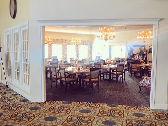 View through a large open doorway into a dining room with multiple tables and chairs arranged neatly. The room is well-lit with natural light coming through large windows with floral curtains. Several chandeliers hang from the ceiling, and the carpet has a dark pattern. The foreground shows a patterned carpet in the adjacent room.