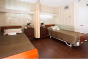 Two single beds with brown bedspreads separated by a privacy curtain, each with a nightstand and lamp in a small, well-lit room.