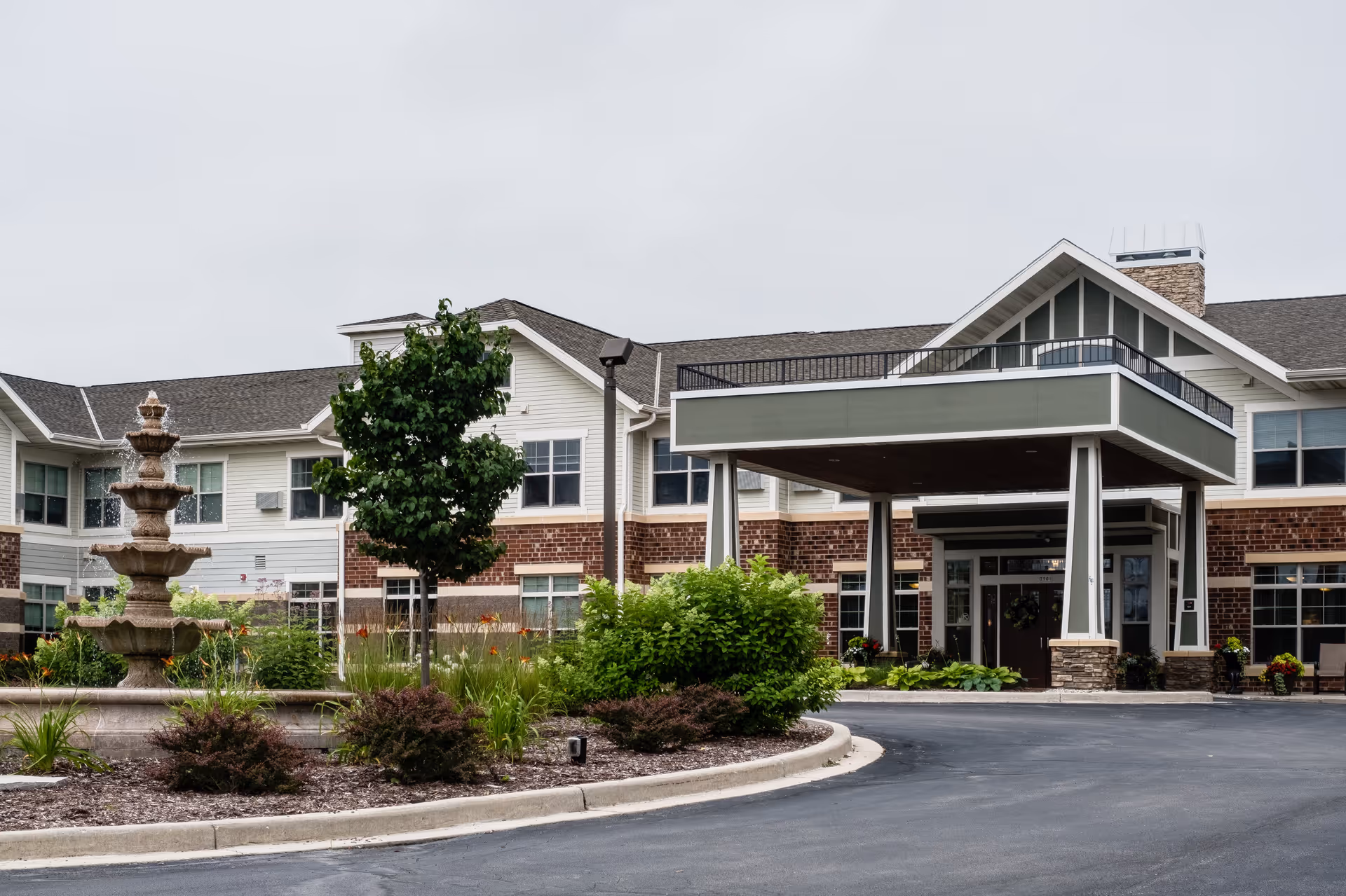 Exterior view of Heritage West Allis senior living facility showing a two-story building with a covered entrance, landscaped garden with a multi-tiered fountain, bushes, and a tree in front.
