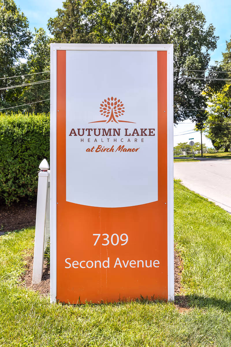 A tall rectangular sign for Autumn Lake Healthcare at Birch Manor, located at 7309 Second Avenue, standing on a grassy area with trees and a road in the background.