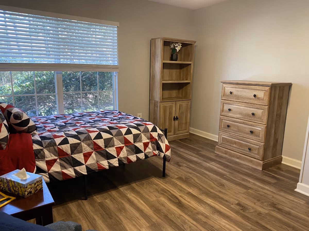 A bedroom with a bed covered in a geometric patterned quilt in red, black, gray, and white. There is a wooden dresser with four drawers and a matching wooden bookshelf with a small vase of flowers on the top shelf. The room has wood flooring and a large window with blinds partially open, showing greenery outside.