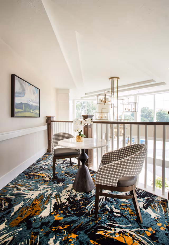 Small seating area with two upholstered chairs and a round marble table overlooking a bright atrium with hanging light fixtures.