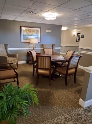 A cozy seating area in a senior living facility with a round wooden table surrounded by six upholstered chairs. Two wingback chairs are positioned against the wall, which is decorated with a framed landscape painting and a table lamp. The room has beige walls with white trim, carpeted floors, and ceiling lights. A green potted plant is visible in the foreground.