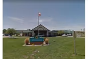 Exterior view of The Meadows senior living facility building with a green lawn in front, an American flag on a flagpole, and a sign that reads 'Meadows'. The sky is clear with a few clouds.
