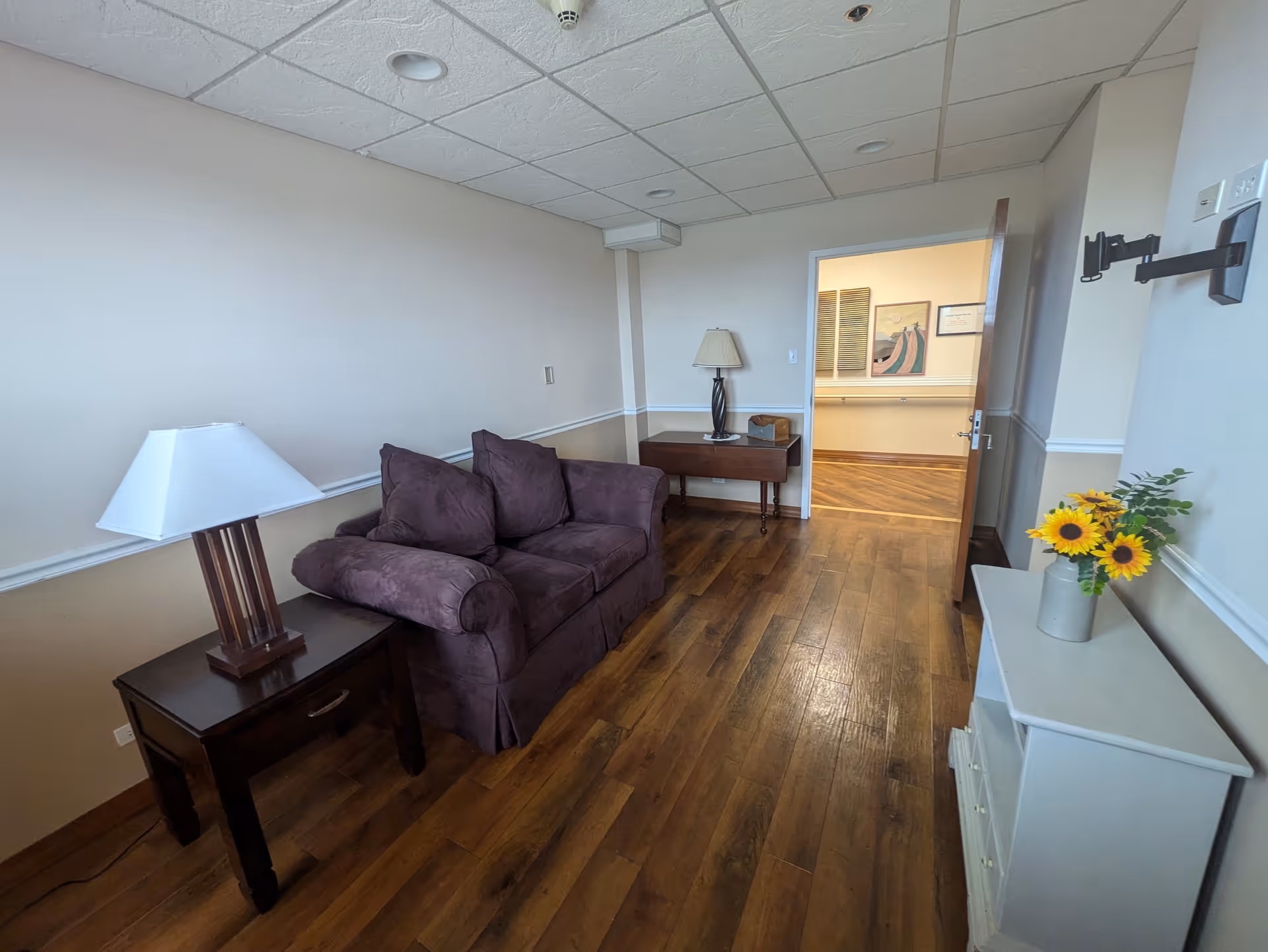 A small living room with a purple couch, a wooden side table with a lamp, a wooden floor, and a white cabinet with a vase of sunflowers. The room has beige walls with white trim and a ceiling with recessed lights. An open door leads to a hallway with framed artwork on the walls.