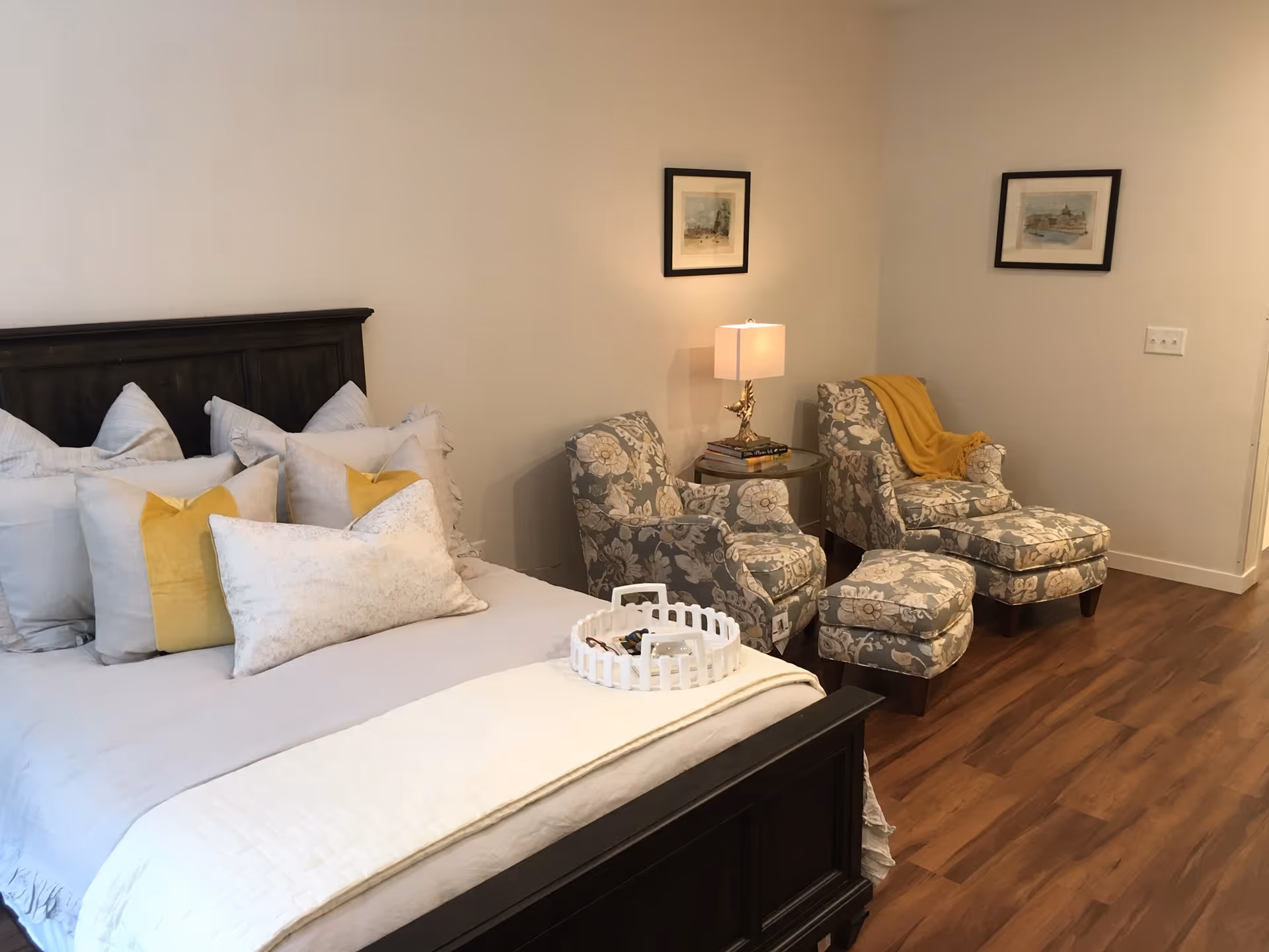 Bedroom with a dark wood bed dressed in layered pillows and a pair of patterned armchairs with ottoman and a side table lamp on hardwood flooring.