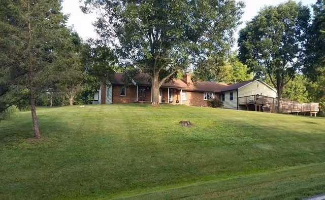 A single-story brick and siding building situated on a grassy hill with several large trees surrounding it. The building has a porch area and a wooden deck extending from one side. The sky is clear and the scene is well-lit by natural daylight.