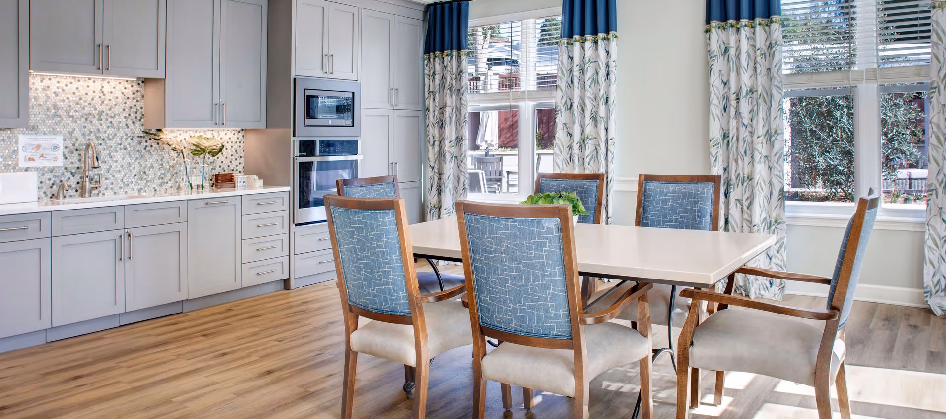 Sunlit dining area with a table and six upholstered chairs beside a modern kitchenette and large windows with patterned curtains.