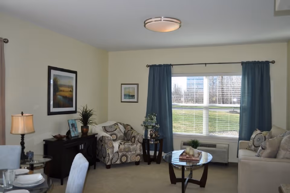 Bright furnished living room with a sofa, patterned armchair, coffee table, sideboard and a large window with blue curtains.
