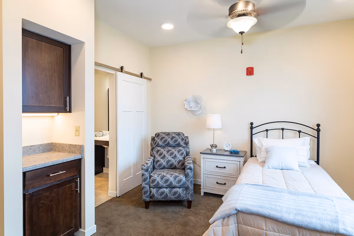A cozy bedroom in an assisted living facility featuring a single bed with white bedding and pillows, a gray nightstand with a lamp, clock, and decorative items, a patterned armchair, a ceiling fan with light, and a sliding barn door leading to a bathroom. There is also a small kitchenette area with dark wood cabinets and a countertop.