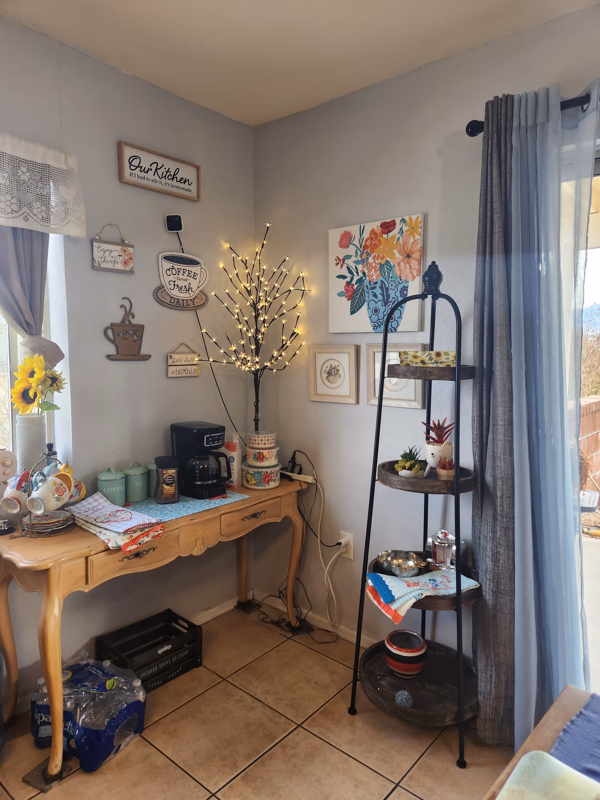 A cozy kitchen corner with a wooden table holding a coffee maker, coffee canisters, and decorative items. The wall above the table is decorated with various signs and framed artwork, including a sign that says 'Our Kitchen' and a coffee-themed sign. A small tree with string lights stands on the table. Next to the table is a tall, black metal shelf with three wooden tiers holding small plants and decorative items. A window with lace curtains and a sliding glass door with gray curtains are visible, letting in natural light.