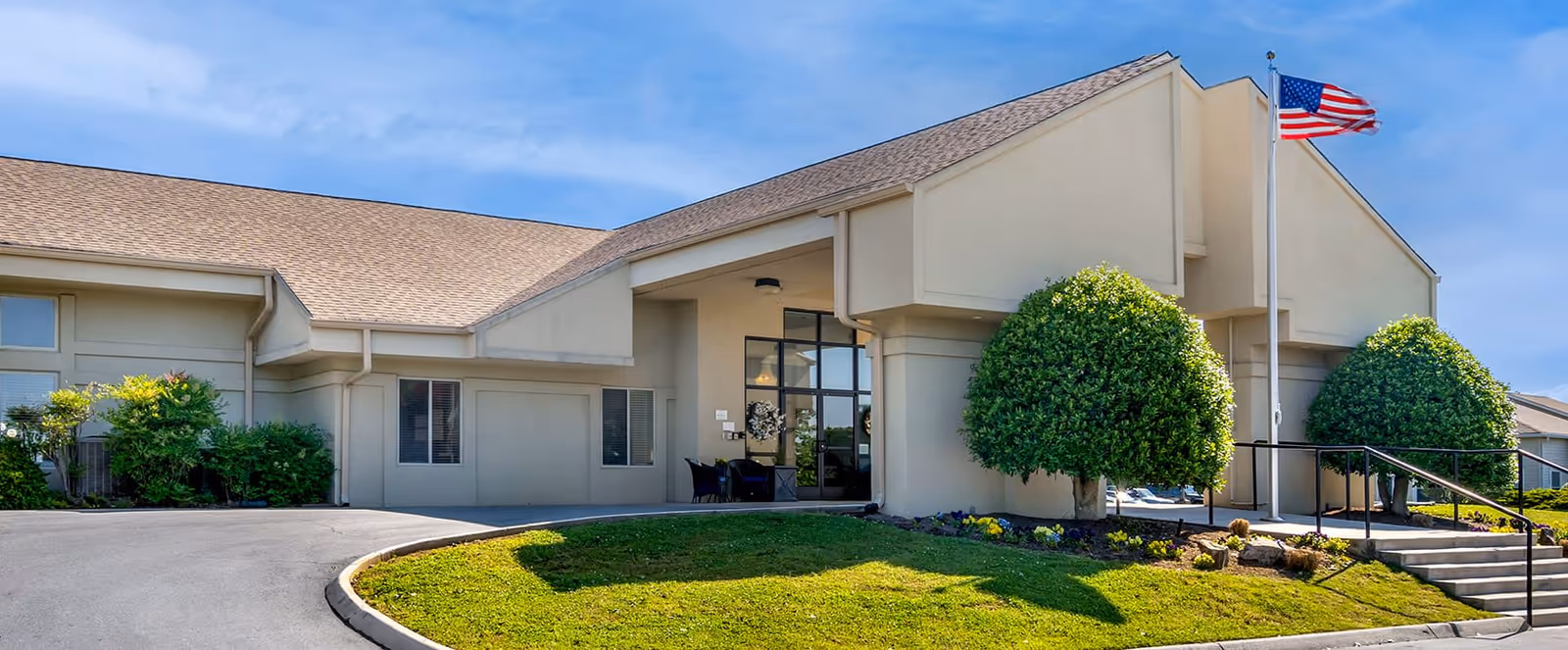 Exterior view of a single-story building with beige walls and a tan roof, featuring a main entrance with glass doors and windows. There are two neatly trimmed round bushes near the entrance, a small garden area with flowers, a flagpole with an American flag, and a paved driveway leading up to the building under a blue sky with some clouds.
