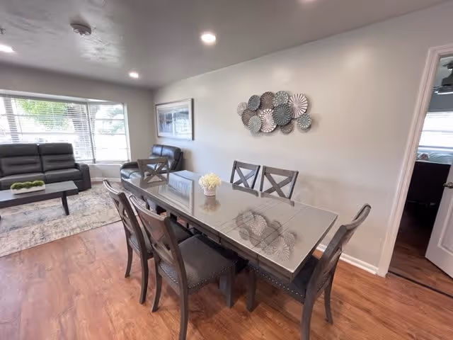 Glass-topped dining table with six chairs in an open-plan room leading to a living area with sofas and a large window.