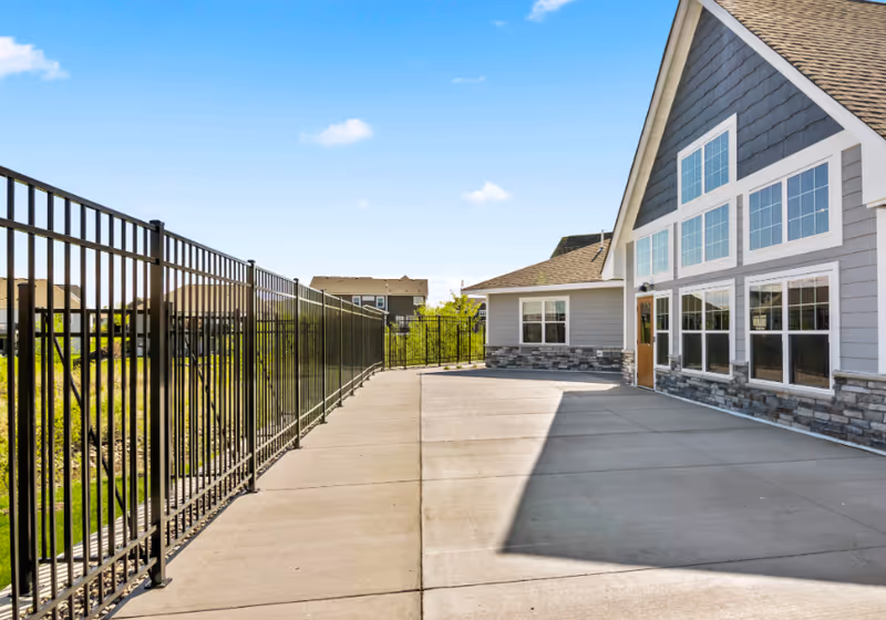 Paved outdoor patio beside a senior living building with large windows, stone siding, and a metal fence under a blue sky.