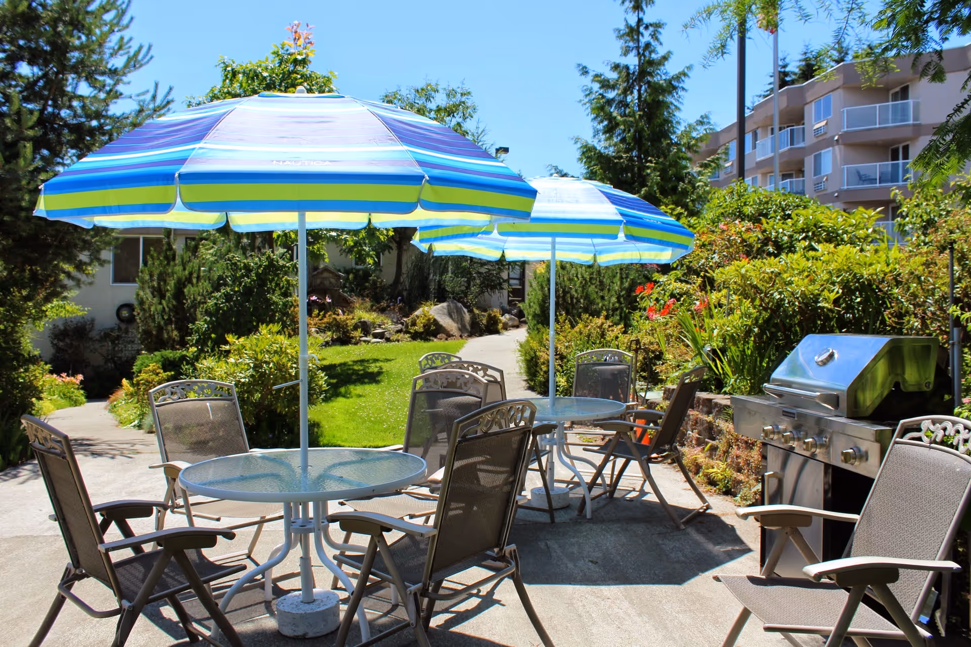 Outdoor patio area with two round glass tables, each shaded by a blue and green striped umbrella. Several metal mesh chairs surround the tables. A stainless steel barbecue grill is visible on the right side. The area is surrounded by green bushes, trees, and a multi-story building in the background under a clear blue sky.