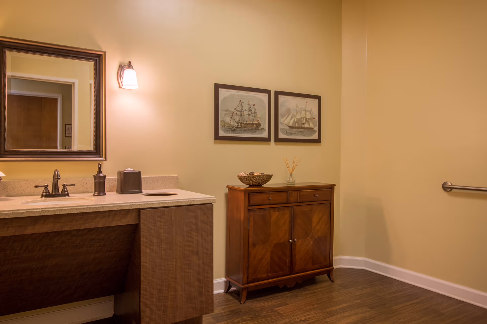 Well-lit bathroom with a sink and mirror on the left and a wooden cabinet topped with decor and framed ship prints on the right.