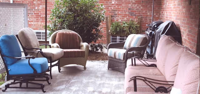 Covered outdoor patio with cushioned wicker and metal seating arranged around potted plants and a grill against a brick wall.