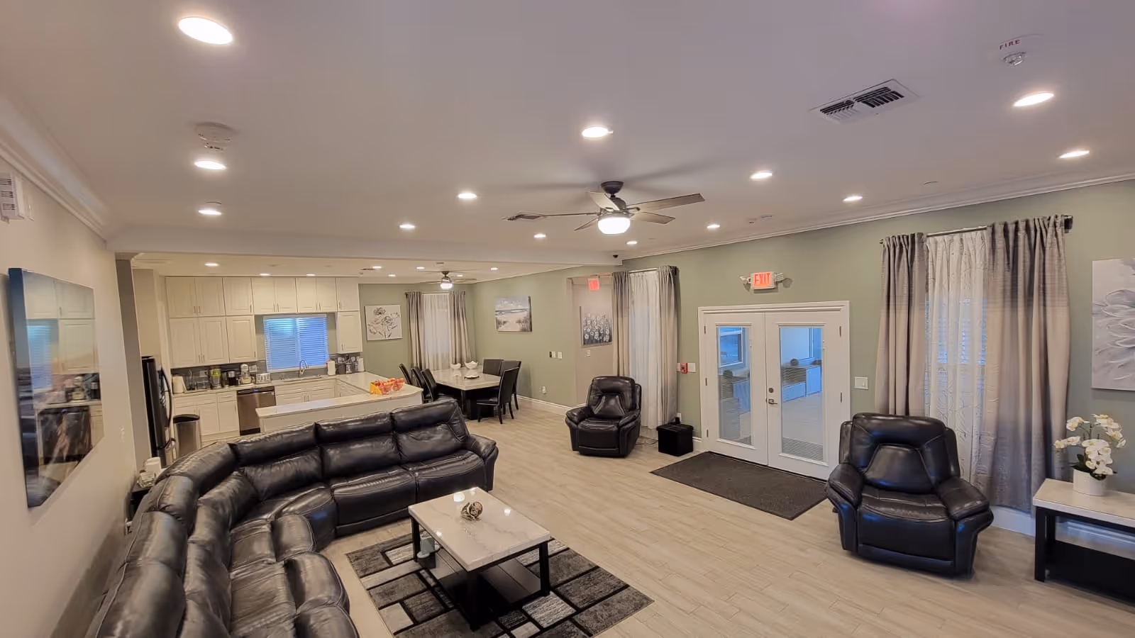 A spacious, well-lit common area in an assisted living facility featuring a large black leather sectional sofa, two black leather armchairs, a coffee table on a patterned rug, and a dining table with chairs near a kitchen area with white cabinetry and appliances. The room has light wood flooring, green walls, ceiling fans, and large windows with curtains.