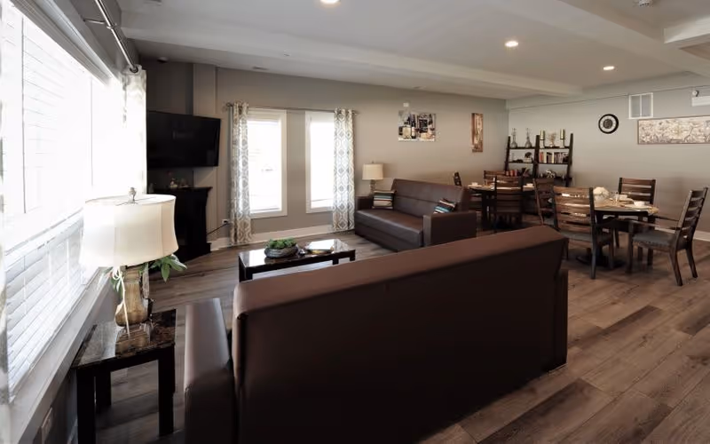 A bright and spacious living room area with two brown sofas, a coffee table, a side table with a lamp, a wall-mounted TV, and large windows with curtains letting in natural light. In the background, there is a dining area with multiple wooden tables and chairs, a shelving unit with decorative items, and wall art.