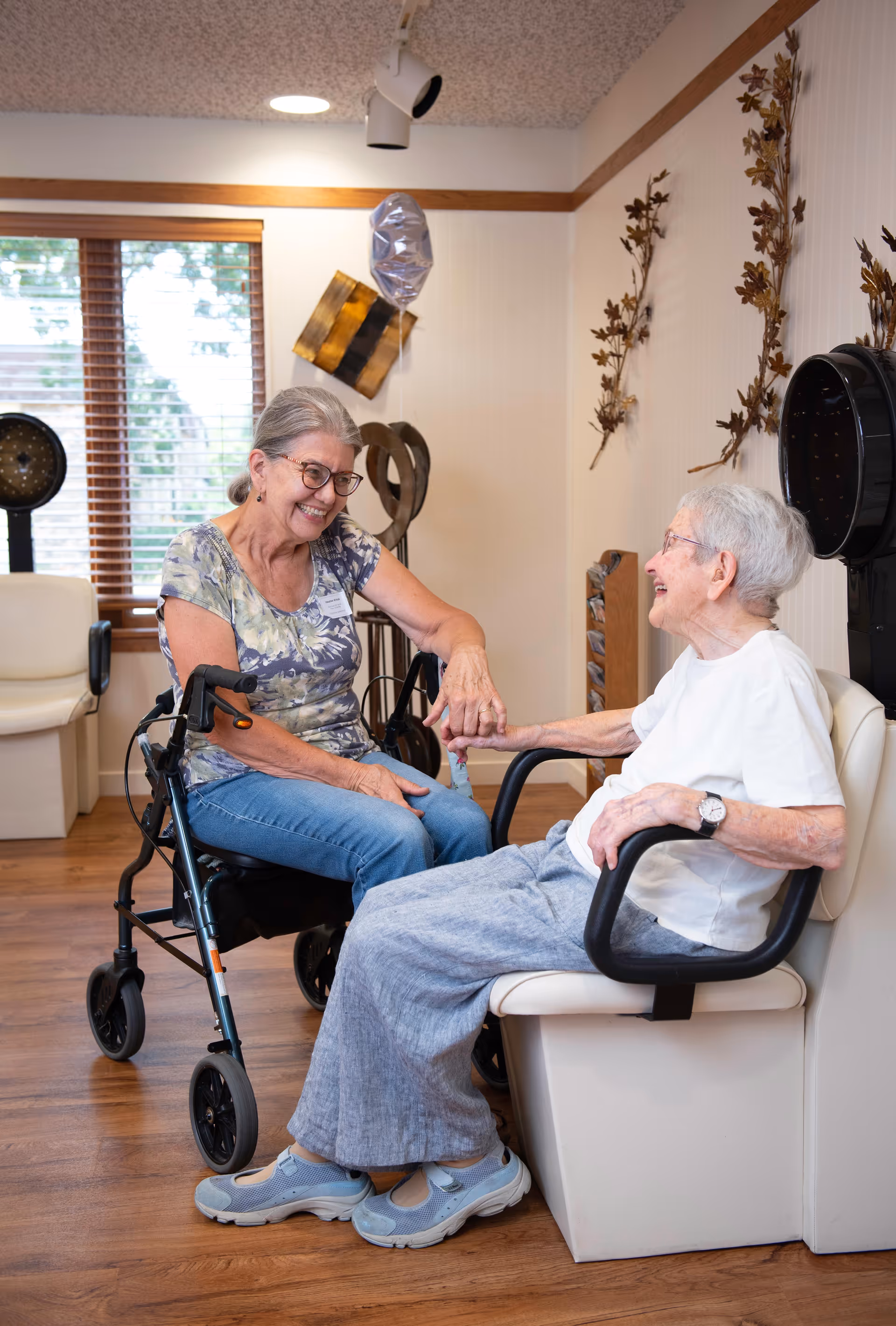 Two elderly women smiling and holding hands in a cozy room with wooden floors and decorative wall art. One woman is seated in a wheelchair, and the other is sitting in a salon chair under a hair dryer. A window with blinds is visible in the background.