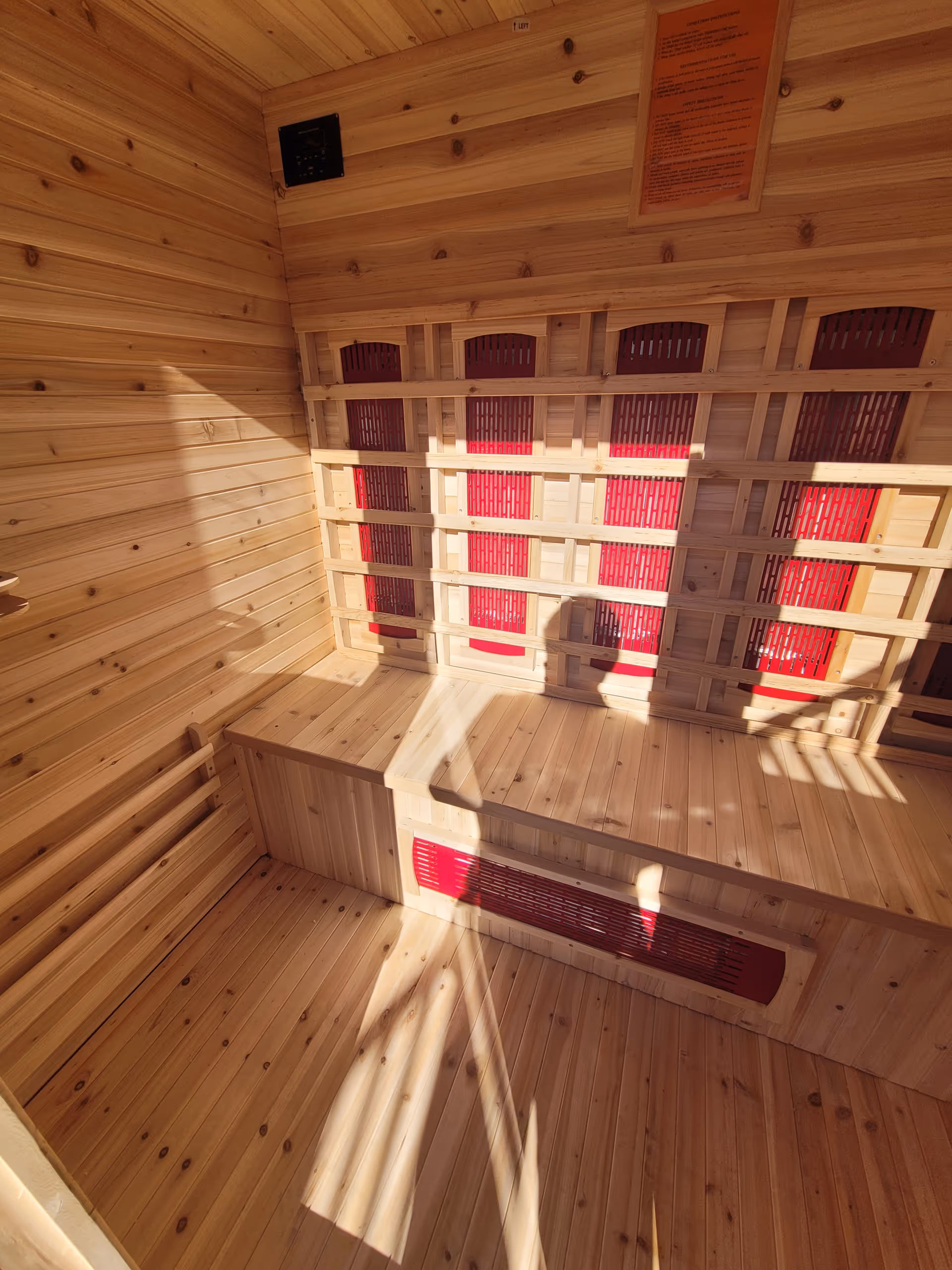 Interior of a wooden sauna room with wooden benches and red heating panels on the walls. Sunlight casts shadows inside the room, including the shadow of a person taking the photo.