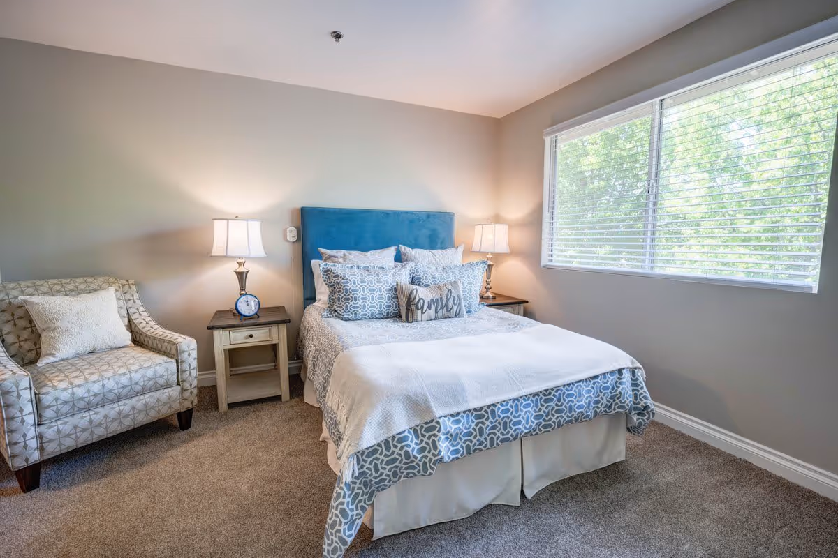 A cozy bedroom with a bed featuring a blue upholstered headboard, multiple patterned pillows including one with the word 'family', and a light blue and white bedspread. There are two wooden nightstands on either side of the bed, each with a lamp. To the left of the bed is a patterned armchair with a white pillow. A large window with white blinds allows natural light to fill the room, and the walls are painted a soft beige color.