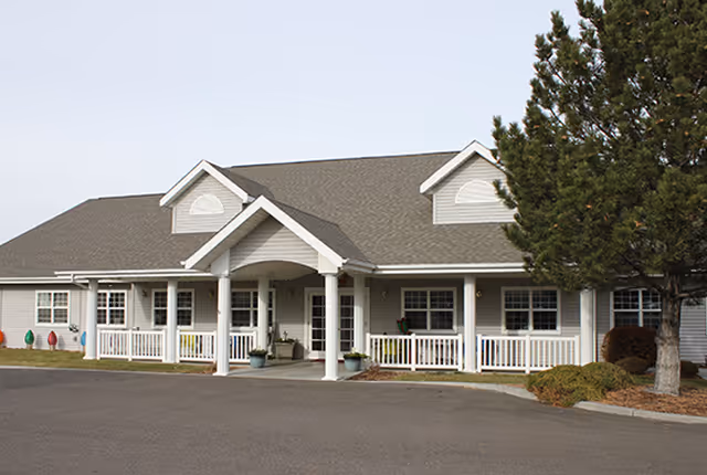 Exterior view of a single-story senior living facility building with a covered entrance supported by white columns, multiple windows, a gray roof, and a tree on the right side.
