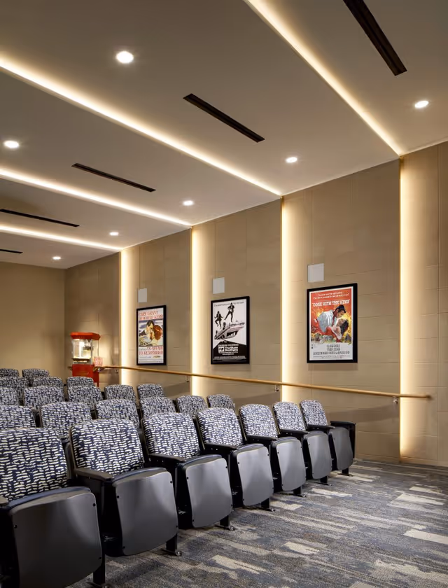 Small theater screening room with rows of patterned upholstered seats, framed movie posters on the wall, and a popcorn machine in the corner.