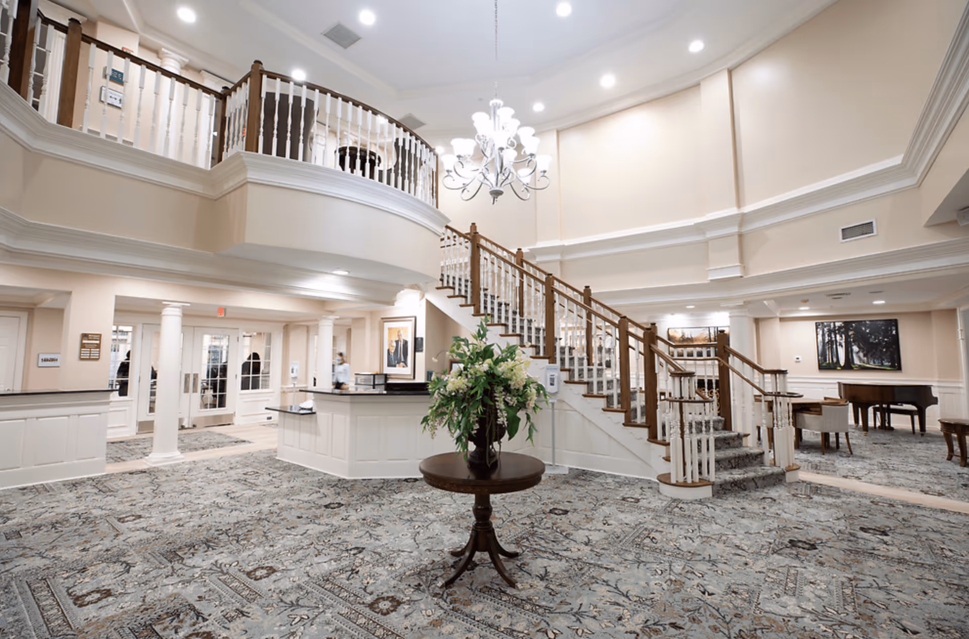 Spacious and elegant interior lobby area of a senior living facility with a central round table holding a floral arrangement, a grand staircase with wooden handrails leading to an upper floor balcony, a chandelier hanging from the ceiling, and a seating area with a piano and chairs in the background.